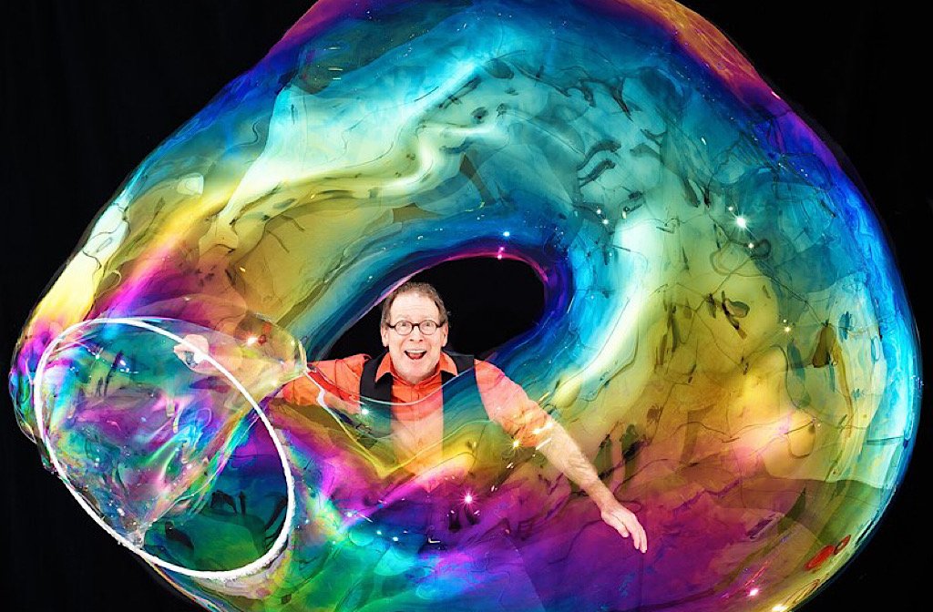 A man in a pink shirt and black waistcoat stands inside a huge swirly bubble that he has created