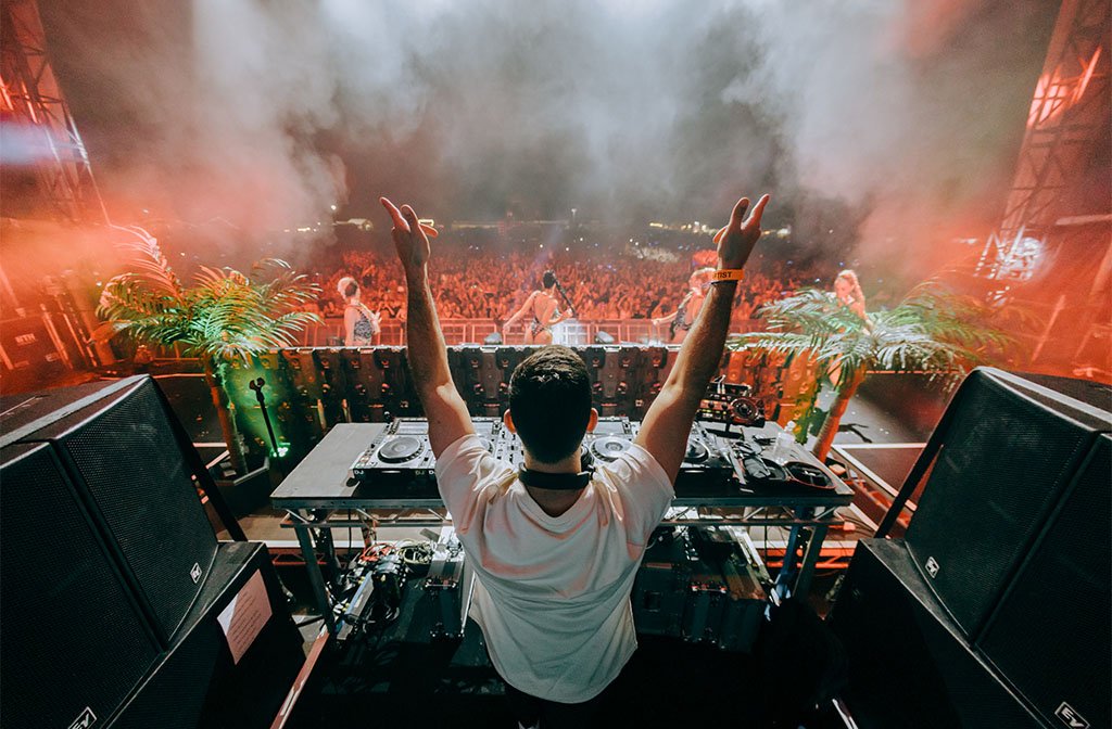 A DJ pictured from behind holding his arms in the air in front of a crowd in Bedford Park. You can see his decks and dancing girls on the stage too. It is nighttime and there is smoke and lots of colourful lights.