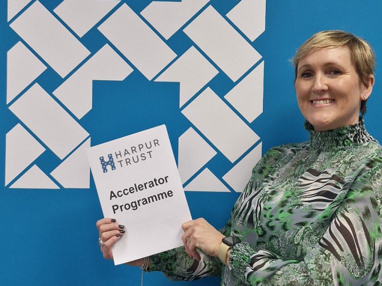 Caroline Downing of the Harpur Trust stands in front of the charity's logo and holds a copy of the Harpur Trust's Accelorator Programme.