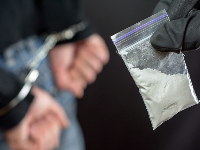 The hand of a police officer holding a small bag of white powder. In the background is a person with their hands behind their back and in handcuffs.