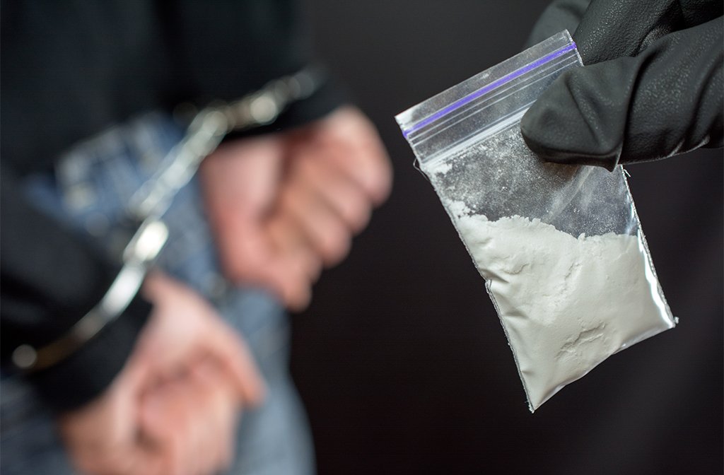 The hand of a police officer holding a small bag of white powder. In the background is a person with their hands behind their back and in handcuffs.