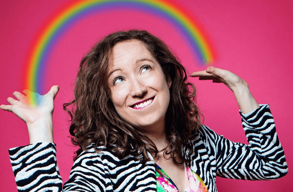 Kaye Leideskope is wearing a zebra print jacket against a pink background and looks like she is holding up a rainbow with both hands