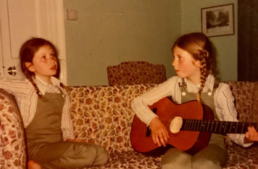 An old family photo of two girls sitting on a patterned sofa wearing identical outfits. One girl is playing a guitar