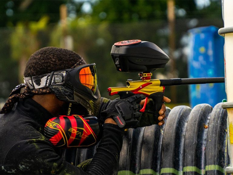 A teenager in goggles and protective gear with a paintball rifle, taking aim at unseen 'enemies' from behind steel oil drums.