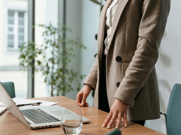 Person in Brown Jacket standing at an office desk with one hand on the table. Image: Mart Production/Pexels