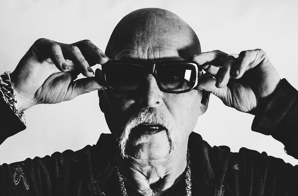 A black and white photo of Phil Hartnoll of Orbital. He is wearing sun glasses and has a statement moustache