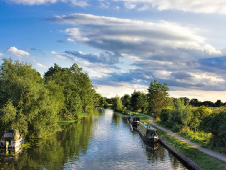 The River Great Ouse from above showing a narrow boat moored near a path to the right and trees full of green leaves on the left.