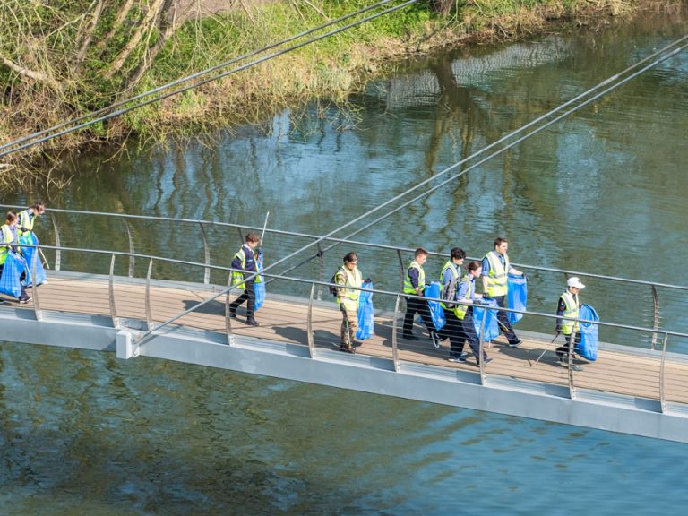 volunteers carrying blue sacks and litterpicking equipment are walking across a bridge in Bedford as part of the Rivercare Clean Up event