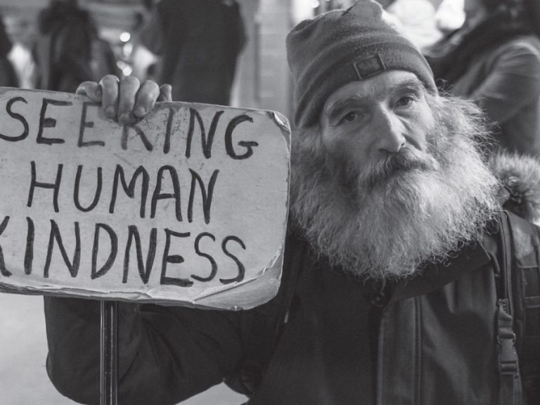 A black and white photo of a homeless man with a big beard wearing a hat and holding up a sign that says "Seeking human kindness"