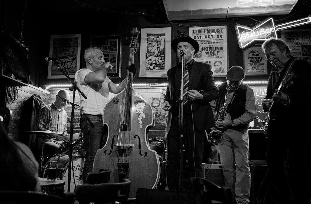 A black and white photo of the band The 251s performing on a small stage. The lead singer is wearing a trilby hat and one band member has a double bass