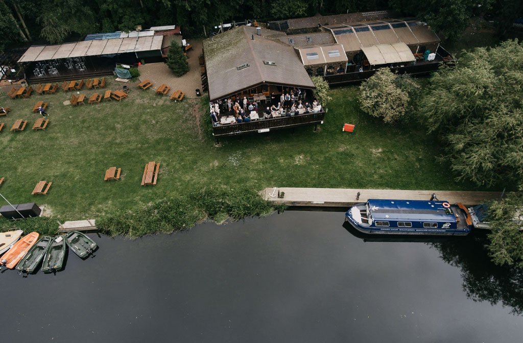 An aerial view of the beautiful riverside setting of the Danish Camp. The image shows the cabin style cafe with a veranda of outdoor seating, plus picnic tables on the lawn overlooking the River Great Ouse and a canal boat on the mooring deck. image Dan Davies 2-D Photography