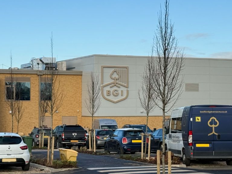 The new look Bedford Greenacre school with cars and a school minibus parked in the foreground. The schools shield with the letters B G and I is on one of the buildings exterior walls.