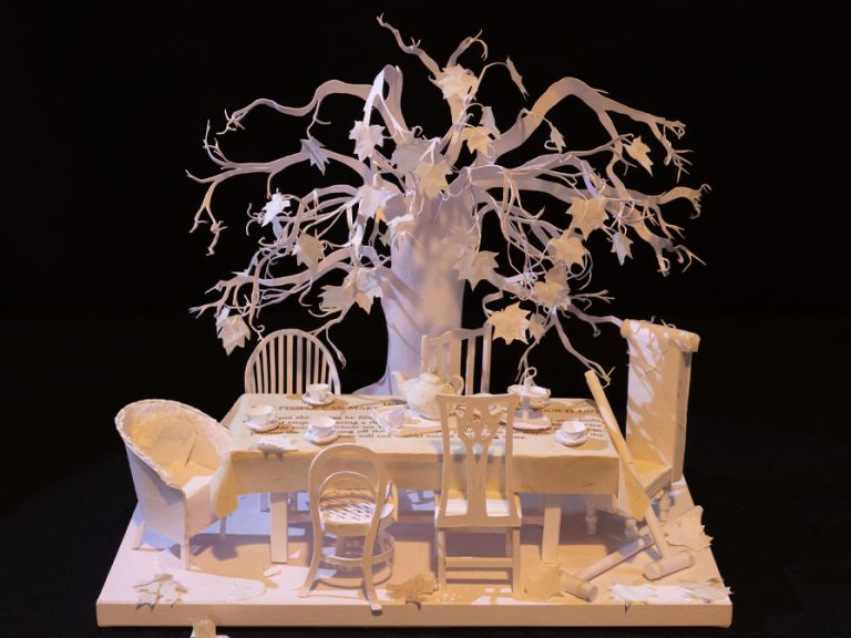 An intricate paper cut model of a dining table surrounded by chair with an autumnal tree in the background. The model is handmade by Jacqui Saunders Love out of white paper and is one a black background