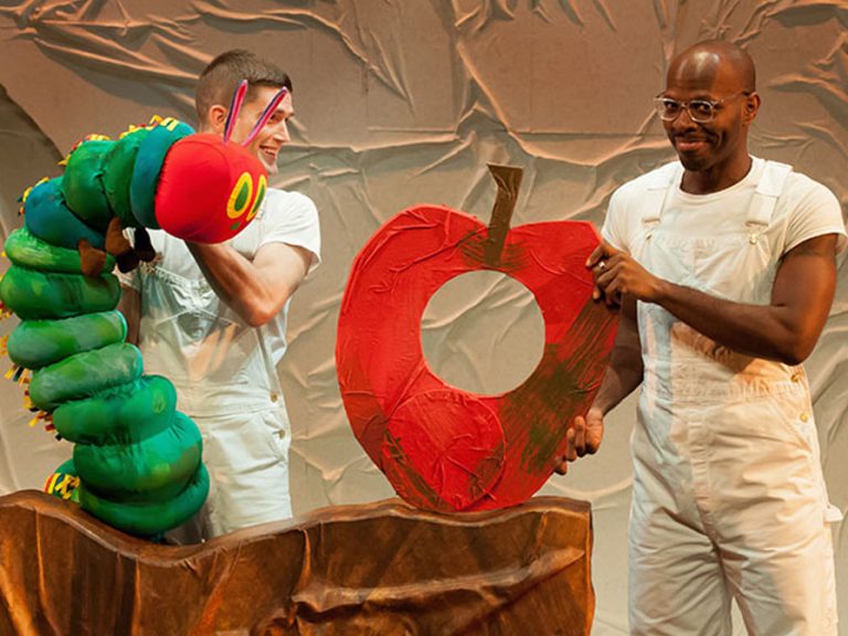 Two puppeteers dressed in white t shirts and trousers are holding puppets of the Very Hungry Caterpillar and an apple with a bite taken out of it