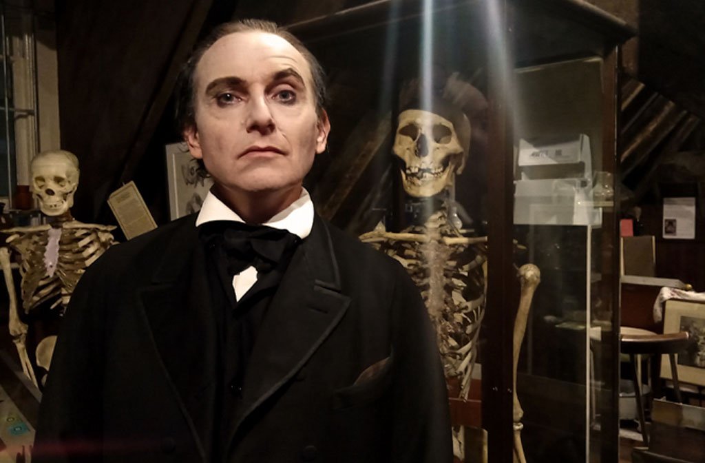 A man is eerily dressed in austere Victorian clothing and surrounded by skeltons