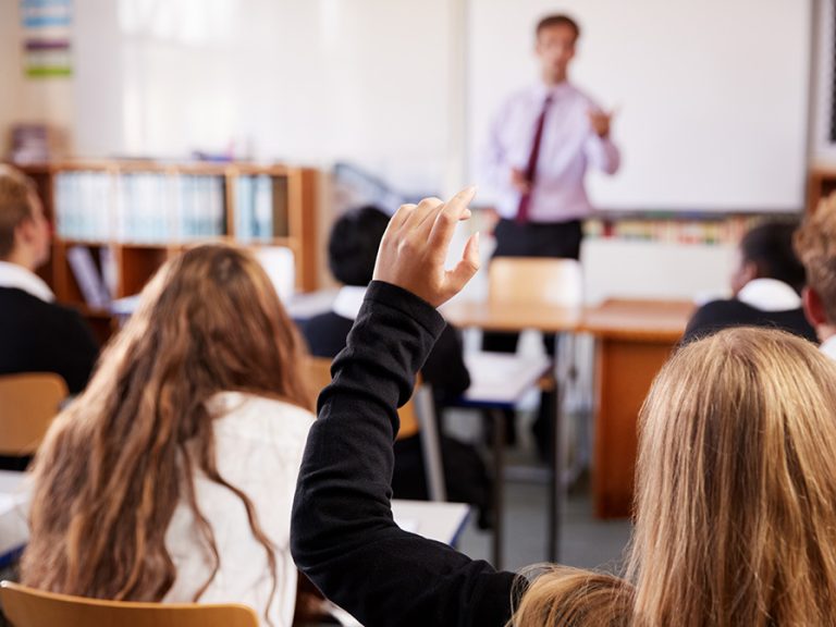 A female pupil raising their hand in a full classroom with the teacher in the background.
