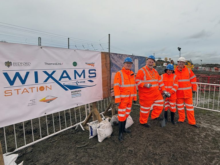 (l-r) Cllr Coombes, Mayor Wootton, Cllr Spice and Cllr Frost all in bright orange hi-vis clothing and hard hats at the site of the new Wixams station. Cllr Slice is holding a shovel and they're standing next to a sign that says 'Wixams'. There are diggers in the background moving earth. Image: Bedford Independent