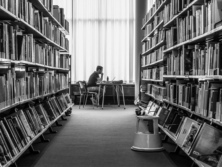A man sits at a table at the end of a long corrridor between two tall shelves filled with books at a library. Image: wal_172619/pixabay