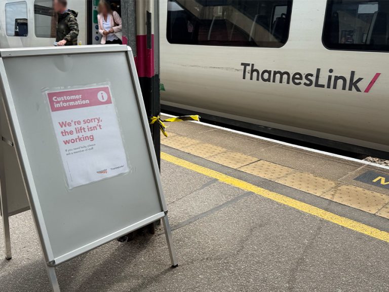 A sign warning train users that the lift at Bedford Train Station is out of order. Passengers are getting off a Thameslink train in the background.