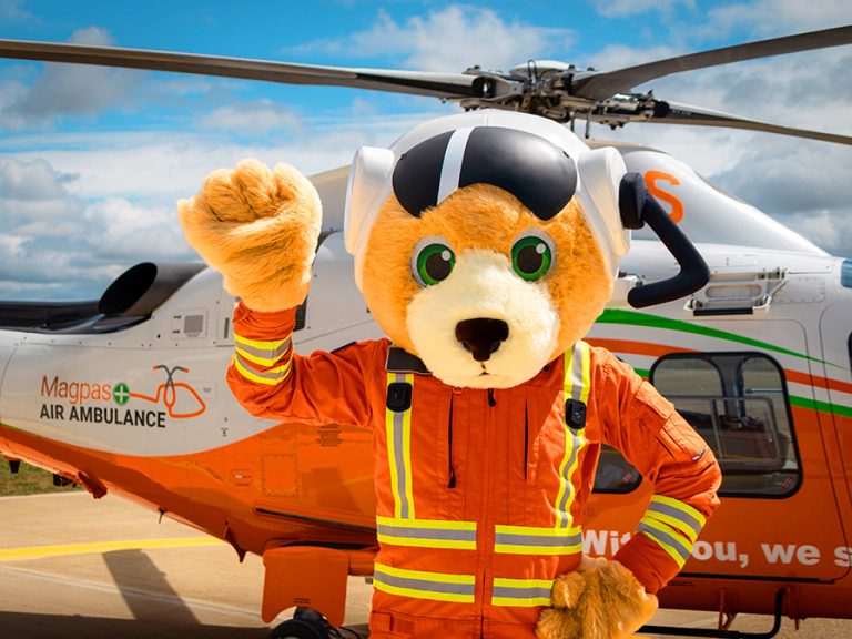 The new Magpas Air Ambulance mascot, a teddy bear in a flight suit, in front of a Magpas helicopter. Image: Magpas
