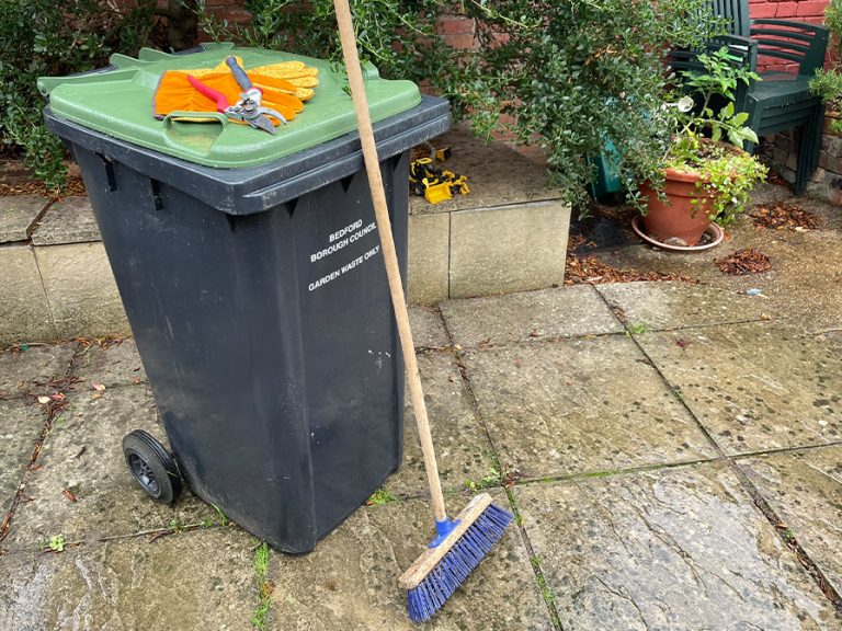 Green wheelie bin in a garden with a broom and gardening equipment Image Bedford Borough Council