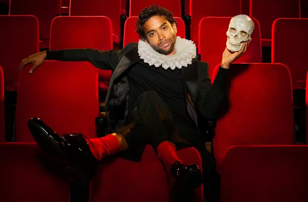 Hiran Abeysekera is pictured as Hamlet in Elizabethan dress wearing a ruff and sitting in a modern theatre holding a skull.