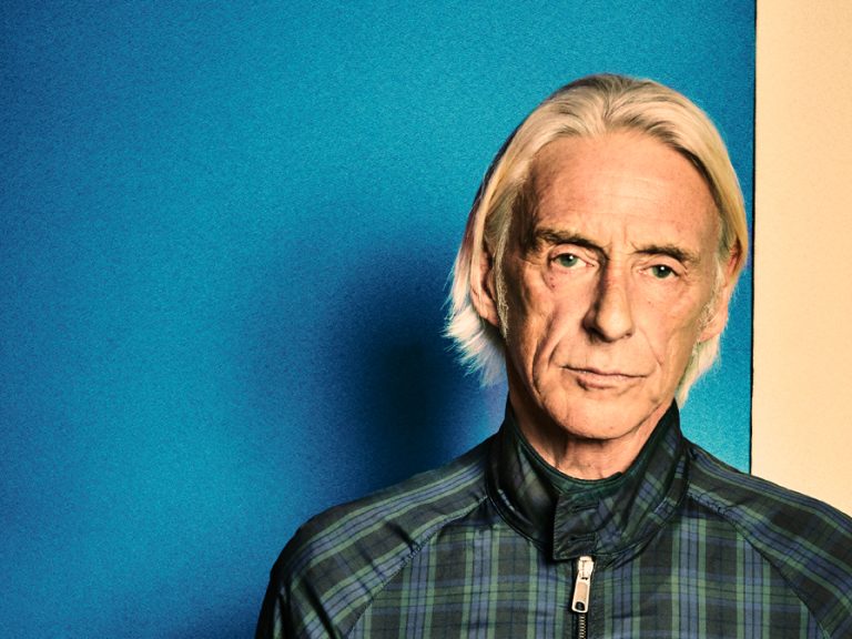 The singer songwriter Paul Weller pictured wearing a checked Harrington jacket against a blue background