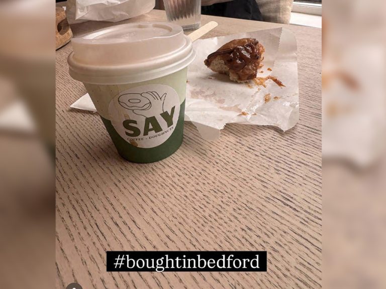A photograph of a takeaway coffee cup and the remains of a doughnut at Say Doughnuts, tagged #BoughtInBedford