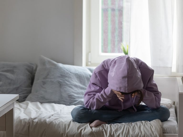 Young teenage girl sitting on bed, covering head with hand Image fizkes Shutterstock