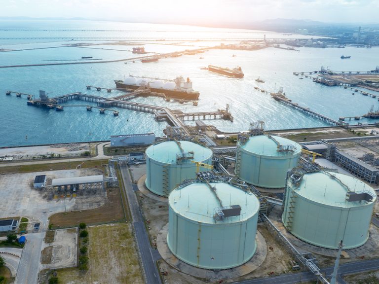 LNG (Liquified Natural Gas) tanker anchored at gas terminal with storage tanks in the foreground and the ocean in the background Image GreenOak Shutterstock