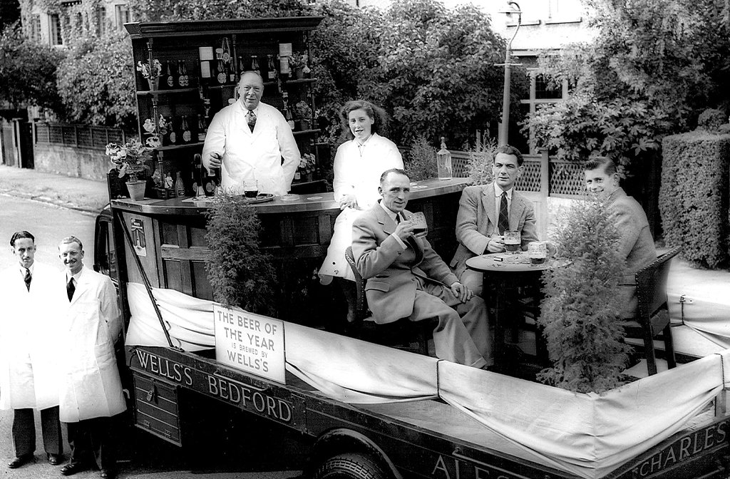 A black and white photo of a mobile bar on the back of a lorry with 'regulars' sat at tables and a bar tender serving Wells beers.