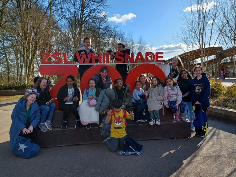 Young carers from Bedfordshire on a trip to Whipsnade Zoo sitting around the entrance sign to the zoo on a bright spring day. Image Carers in Bedfordshire