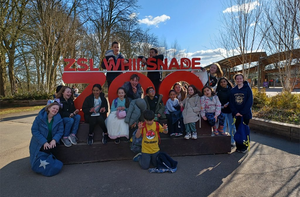 Young carers from Bedfordshire on a trip to Whipsnade Zoo sitting around the entrance sign to the zoo on a bright spring day. Image Carers in Bedfordshire