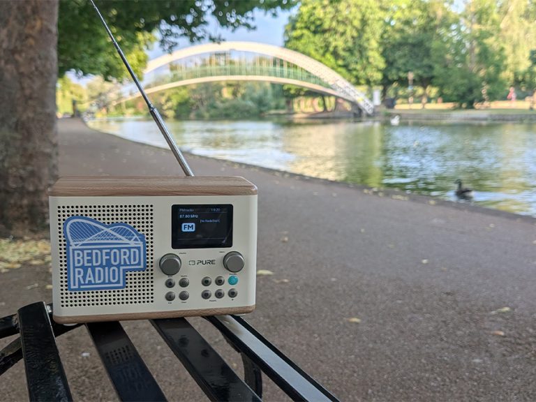 A radio branded with the Bedford Radio logo on a bench by the River Great Ouse and the suspension bridge in the background