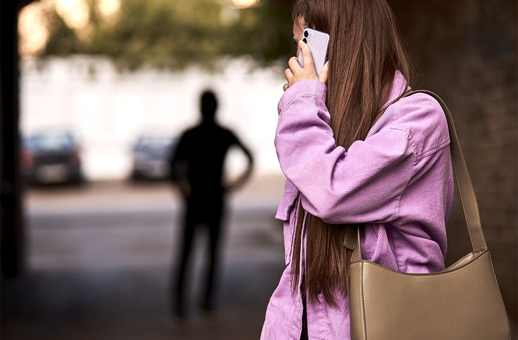 Criminal stalking a woman talking on her mobile phone in dark street, she is looking back at the shadowy figure in the background. Image: Shakirov Albert/Shutterstock