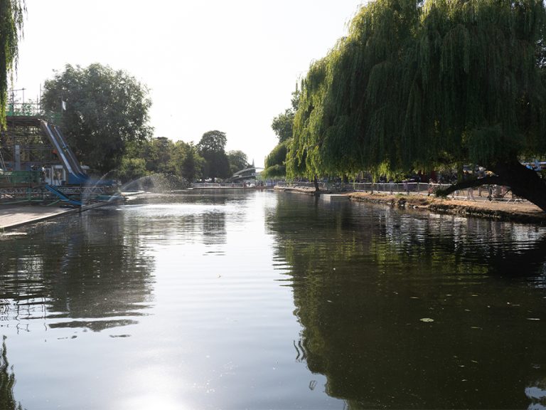 The canoe display area Bedford River Festival 2026. The canoe slides are to the left, with the water of the river still and calm. The suspension bridge can be seen in the background. Willow trees hang over the river on the right.