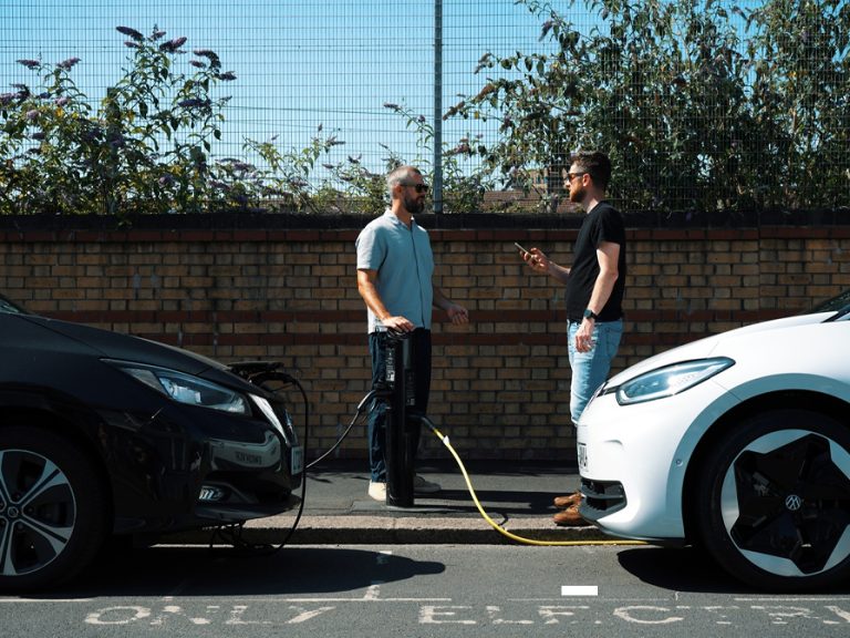 Two men standing and talking at an EV charging point with two cars parked and plugged in. Image: Connected Kerb/Bedford Borough Council.