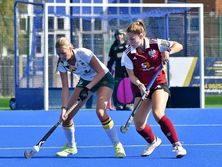 Two hockey players battle for the ball on the blue-clad surface of Chester Road
